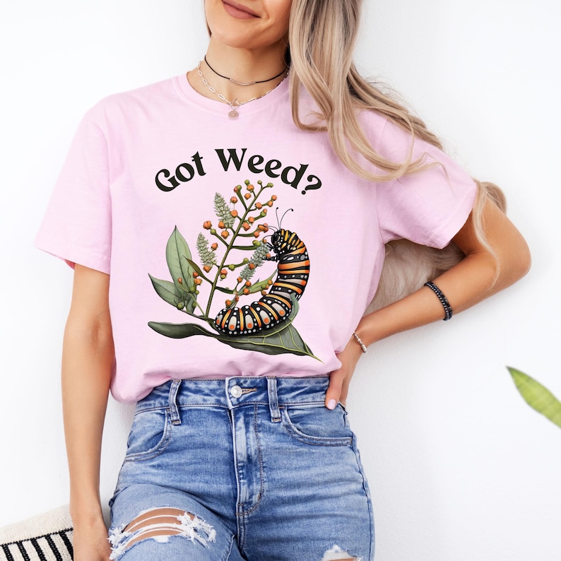 May include: A light pink t-shirt with the text "Got Weed?" in black. The shirt features a colorful illustration of a caterpillar on a plant with green leaves and orange berries. The shirt is made of a soft material.