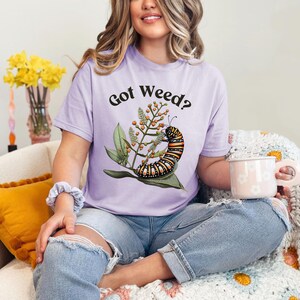 May include: A light purple t-shirt with the text "Got Weed?" above a colorful caterpillar illustration. The caterpillar is on a green leaf with orange berries. The shirt is worn with blue jeans.