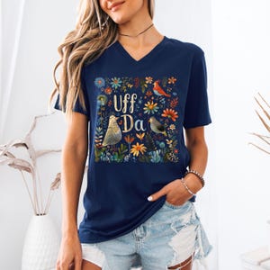 May include: Navy blue V-neck t-shirt featuring a floral and bird design with the text "Uff Da". The design includes colorful flowers and two birds. The shirt is made of a soft material.