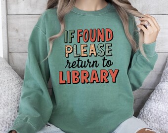 May include: A sage green crewneck sweatshirt featuring the text IF FOUND PLEASE return to LIBRARY in a mix of bold orange, tan, and black lettering. The garment has long sleeves and a ribbed neckline, cuffs, and hem, with a soft, worn-in texture.