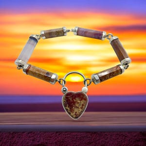 May include: A gemstone bracelet with a heart-shaped clasp. The bracelet features cylindrical beads in shades of brown, pink, and white, connected by silver links. The heart-shaped clasp is a reddish-brown color with a speckled pattern.