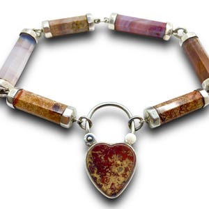 May include: A silver bracelet featuring cylindrical gemstone beads in shades of brown, pink, and purple. The bracelet is secured with a heart-shaped lock. The gemstones are approximately 0.5 inches in length.