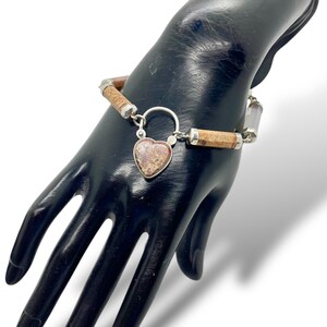 May include: A silver bracelet with a heart-shaped pendant. The bracelet features cylindrical brown and silver beads. The heart pendant has a speckled, reddish-brown appearance. The bracelet is displayed on a black hand.