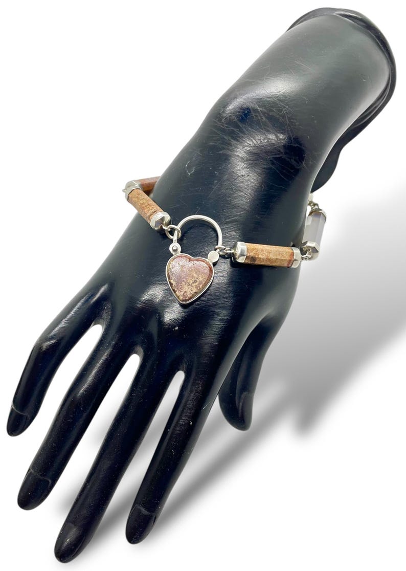 May include: A silver bracelet with a heart-shaped pendant. The bracelet features cylindrical brown and silver beads. The heart pendant has a speckled, reddish-brown appearance. The bracelet is displayed on a black hand.