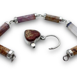 May include: A gemstone bracelet with a heart-shaped clasp. The bracelet features cylindrical beads in shades of brown, purple, and white, connected by silver links. The heart-shaped clasp is a reddish-brown color.