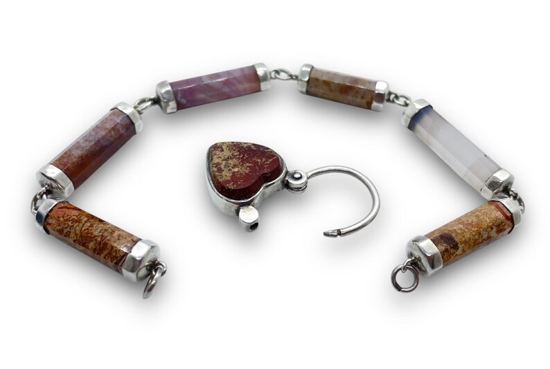 May include: A gemstone bracelet with a heart-shaped clasp. The bracelet features cylindrical beads in shades of brown, purple, and white, connected by silver links. The heart-shaped clasp is a reddish-brown color.