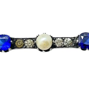 May include: A silver bar brooch with a pearl and two blue gemstones set in a row. The brooch is decorated with smaller clear and gold-toned gemstones.