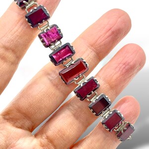May include: A silver bracelet with a row of rectangular red, pink, and purple gemstones. The gemstones are set in silver and have a faceted cut.