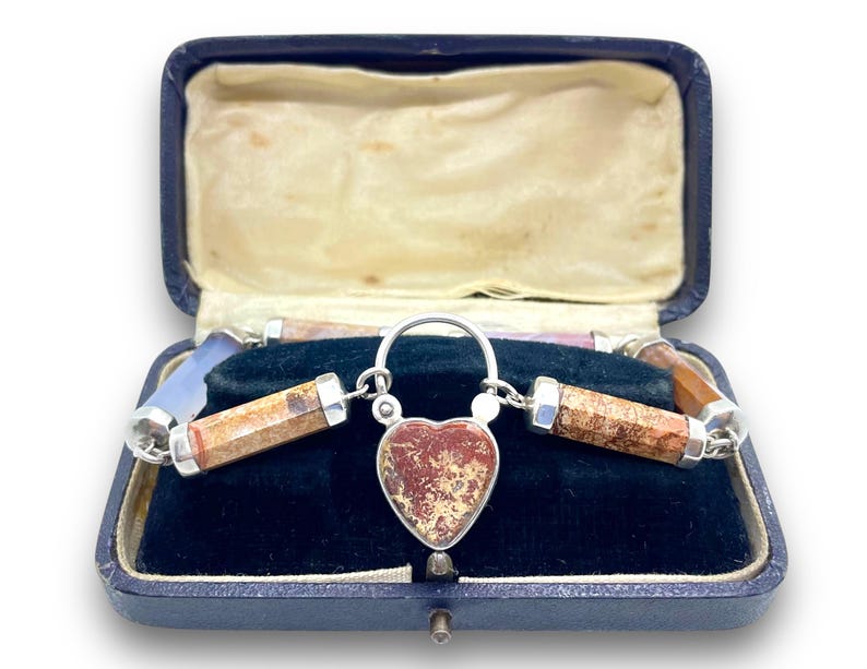 May include: A vintage silver necklace with a heart-shaped pendant and cylindrical stone beads, displayed in a blue velvet-lined box. The pendant and beads feature earthy tones of brown, red, and beige.