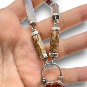 May include: A silver necklace featuring a heart-shaped pendant with red and tan patterns. The necklace has cylindrical stone beads in shades of tan, brown, and lavender, connected by silver links. The pendant is approximately 1 inch wide.