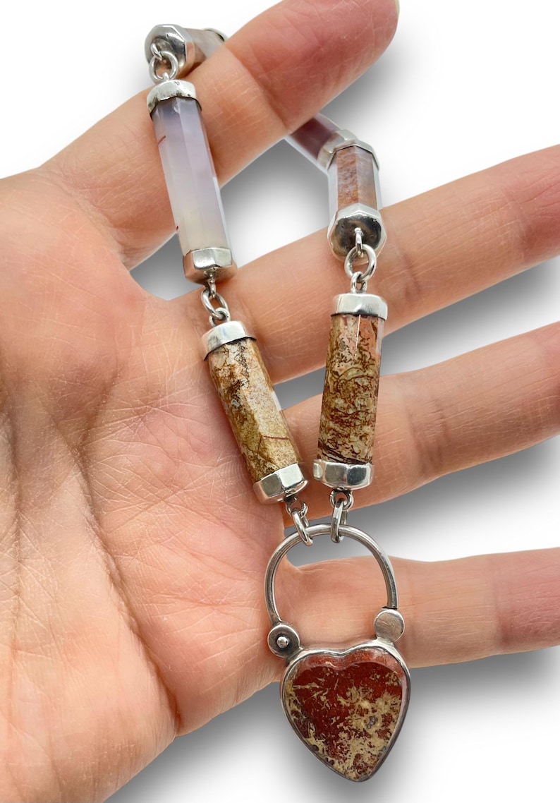 May include: A silver necklace featuring a heart-shaped pendant with red and tan patterns. The necklace has cylindrical stone beads in shades of tan, brown, and lavender, connected by silver links. The pendant is approximately 1 inch wide.