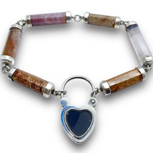 May include: A gemstone bracelet featuring cylindrical beads in shades of brown, purple, and white, connected by silver links. The bracelet includes a heart-shaped lock with a dark blue center, adding a unique touch to this jewelry piece.