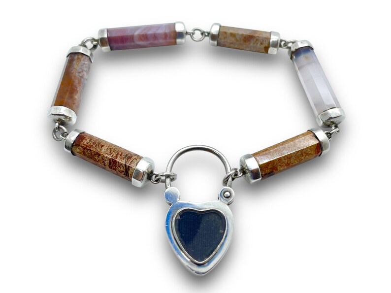 May include: A gemstone bracelet featuring cylindrical beads in shades of brown, purple, and white, connected by silver links. The bracelet includes a heart-shaped lock with a dark blue center, adding a unique touch to this jewelry piece.