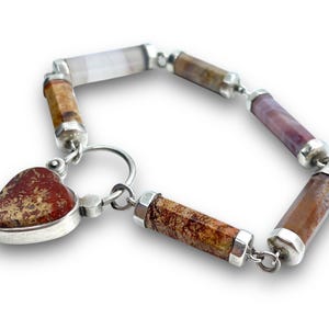 May include: A gemstone bracelet featuring a heart-shaped pendant and cylindrical beads in shades of brown, purple, and white. The bracelet is connected with silver links and has a silver clasp. The heart pendant has a speckled pattern.