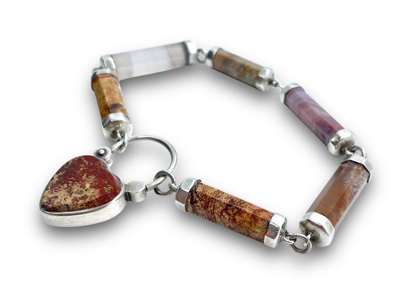 May include: A gemstone bracelet featuring a heart-shaped pendant and cylindrical beads in shades of brown, purple, and white. The bracelet is connected with silver links and has a silver clasp. The heart pendant has a speckled pattern.