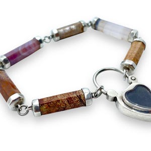 May include: A bracelet featuring cylindrical gemstone beads in shades of brown, red, and purple, connected by silver links. The bracelet includes a heart-shaped charm with a dark blue center. The gemstones are approximately 0.5 inches in length.