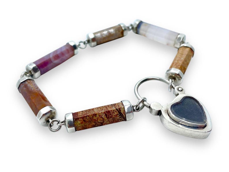 May include: A bracelet featuring cylindrical gemstone beads in shades of brown, red, and purple, connected by silver links. The bracelet includes a heart-shaped charm with a dark blue center. The gemstones are approximately 0.5 inches in length.