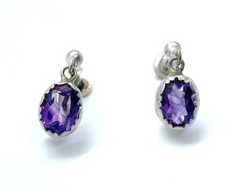 May include: A pair of silver earrings with purple amethyst stones. The earrings have a simple design with a single stone hanging from each earring.