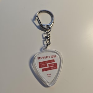 Könnte beinhalten: Ein klarer, gitarrenförmiger Schlüsselanhänger mit einem silbernen Metallverschluss. Der Schlüsselanhänger hat einen weißen Einsatz mit rotem Text, der "BTS WORLD TOUR" und andere Details enthält. Der Schlüsselanhänger ist ein Fan-Accessoire.