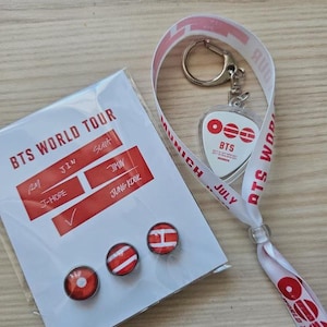 Könnte beinhalten: Ein Set mit BTS World Tour-Merchandise. Es enthält einen transparenten Schlüsselanhänger mit herzförmigem Anhänger, ein Schlüsselband mit dem Text "MUNICH JULY BTS WORLD TOUR" und drei rote und weiße Anstecknadeln. Eine Karte mit den Namen der Bandmitglieder ist ebenfalls enthalten.