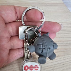 Pode incluir: Um porta-chaves de metal com um pendente de robô esférico cinzento com o número 7.7 em relevo, preso a uma chave prateada e a uma etiqueta de plástico transparente em forma de palheta com logótipos vermelhos, o texto BTS e Munique.