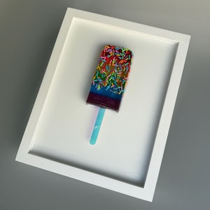 May include: A framed artwork featuring a colorful popsicle with rainbow layers and sprinkles. The popsicle is on a blue stick and is set against a white background.