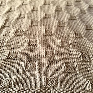 May include: Close-up of a knitted blanket in a warm, neutral brown colour. The blanket features a textured pattern of raised squares and vertical ribbing, creating a cosy and tactile appearance. The bottom edge has a distinct, textured border.
