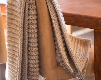 May include: A beige knitted throw blanket draped over a wooden chair. The blanket has a textured, ribbed pattern.