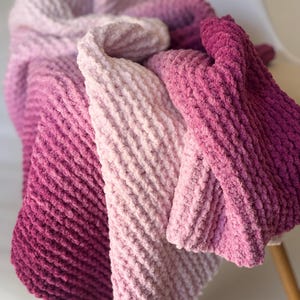 May include: A plush, knitted blanket in shades of pink, from light to deep magenta. The blanket is folded and draped over a chair, showcasing its soft texture and color gradient. The knitted pattern is visible.