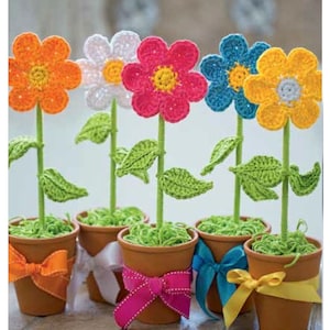 Pode incluir: Seis arranjos de flores de crochê em vasos de terracota com folhas de crochê verdes e laços de fita coloridos. As flores são laranja, branca, rosa, azul e amarela.