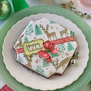 May include: A hexagonal sugar cookie decorated with white icing, featuring a pattern of tan reindeer, green pine trees, and snowflakes. A red icing ribbon crosses the center, topped with a red bow and a yellow plaque reading Happy holidays.