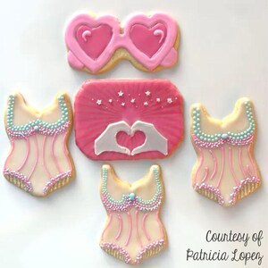 Pode incluir: Cinco biscoitos de a&ccedil;&uacute;car decorados em rosa e branco. Dois biscoitos t&ecirc;m a forma de um cora&ccedil;&atilde;o e s&atilde;o decorados com glac&ecirc; rosa e um contorno branco. Um biscoito tem a forma de um ret&acirc;ngulo e &eacute; decorado com glac&ecirc; rosa e estrelas brancas. Dois biscoitos t&ecirc;m a forma de um corpete e s&atilde;o decorados com glac&ecirc; rosa e branco e granulado azul e branco.