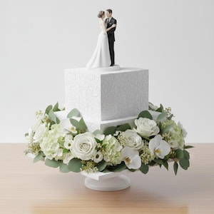 May include: A two-tiered square white cake with a subtle swirling pattern, topped with a bride and groom figurine. The base of the cake is surrounded by a dense arrangement of white roses, white orchids, green hydrangea florets, and eucalyptus leaves.