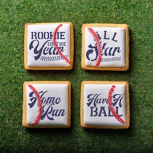May include: Four decorated sugar cookies with baseball-themed designs. The cookies are white with red stitching and black text. The text on the cookies reads "Rookie of the Year", "All Star", "Home Run", and "Have a Ball".