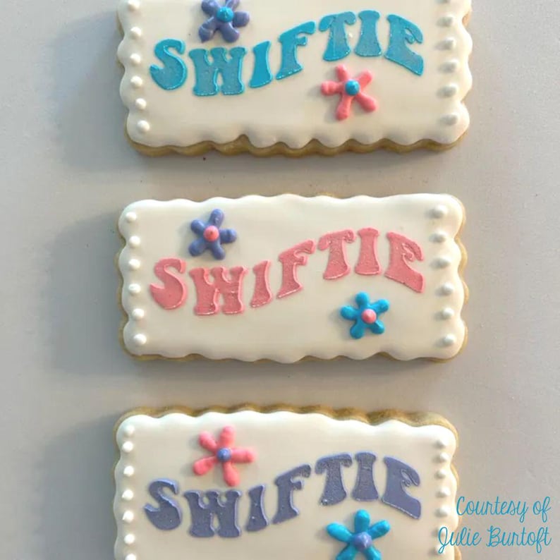 Pode incluir: Tr&ecirc;s biscoitos de a&ccedil;&uacute;car decorados com a palavra "Swiftie" escrita em cores diferentes em cada biscoito. Os biscoitos s&atilde;o decorados com glac&ecirc; branco e flores coloridas.