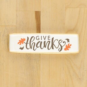 Könnte beinhalten: Ein rechteckiger Keks mit weißer Glasur und dem Schriftzug "give thanks" in brauner Schrift. Orange und braune Blätter zieren den Keks. Er liegt auf einer hellbraunen Holzoberfläche.