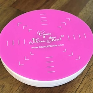 May include: Pink round stencil mat with white markings and the text "Genie Mini-Mat" and "www.StencilGenie.com".