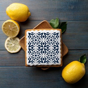 Op de afbeelding: Een witte cake met een blauw en wit geometrisch patroon erop. De cake staat op een houten plank en er liggen citroenen op tafel.