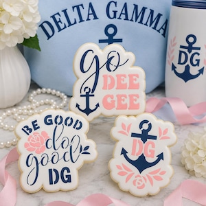 May include: Three white sugar cookies with navy blue and light pink icing. One cookie reads "GO DEE GEE" with an anchor. Another reads "BE GOOD do good DG" with a pink rose. The third features a navy anchor, pink leaves, and "DG" text.
