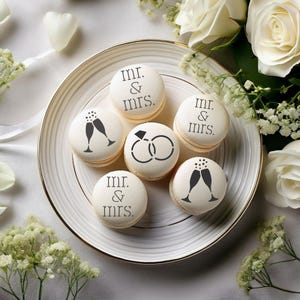 Könnte beinhalten: Sechs Makronen auf einem weißen Teller, verziert mit schwarzen Designs und Text. Einige tragen die Aufschrift "mr. & mrs.", andere zeigen Sektgläser oder Eheringe. Weiße Rosen und Grünzeug bilden eine Hochzeitskulisse.