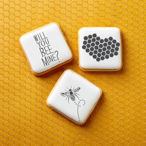 May include: Three white sugar cookies decorated with black icing. One cookie says "Will you bee mine?" Another cookie has a honeycomb design. The third cookie has a black and white bee illustration.