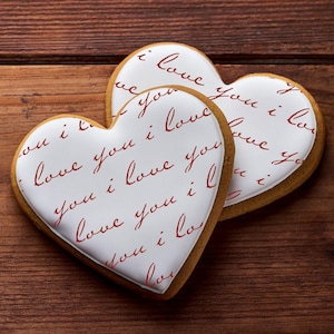 May include: Two heart-shaped cookies with white icing and the words "Love you i love you" in red script. The cookies sit on a dark brown wooden surface, perfect for Valentine's Day or a romantic occasion.