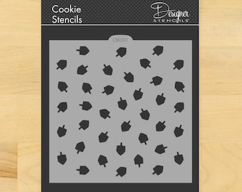 Könnte beinhalten: Eine Keksschablone mit grauem Hintergrund und einem Muster aus schwarzen Dreidels. Die Schablone ist mit "Cookie Stencils" und "Designer Stencils" beschriftet. Die Schablone befindet sich auf einem schwarzen Hintergrund.