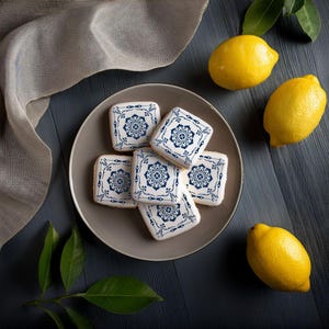Op de afbeelding: Een bord met vierkante koekjes versierd met ingewikkelde blauwe en witte patronen. De koekjes zijn in een cirkel op een grijs bord gerangschikt. Er liggen drie citroenen op een donker houten oppervlak.