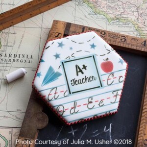 Puede incluir: Una galleta hexagonal decorada con un diseño escolar azul y blanco. La galleta presenta una manzana roja, un avión de papel azul y un rectángulo blanco con el texto "A+ Teacher".