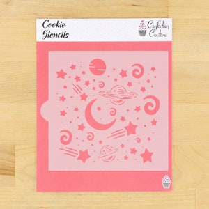 May include: Pink plastic cookie stencil with a space theme. The stencil features planets, stars, shooting stars, and swirls. The text "Cookie Stencils" is at the top of the stencil. The text "Confection Couture" is at the top right of the stencil.