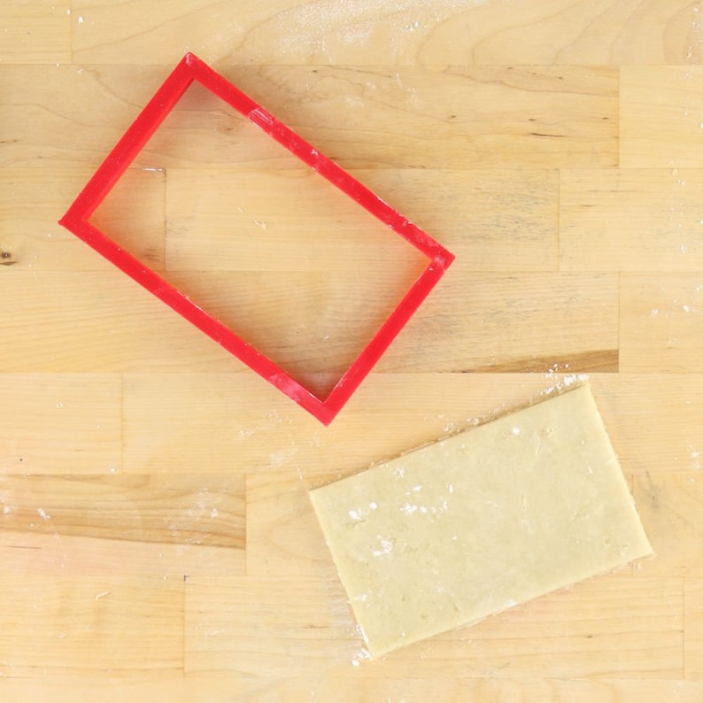 May include: A red plastic rectangle cookie cutter on a wooden surface with a sheet of dough next to it.