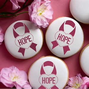 May include: Four white cookies decorated with pink ribbon and heart designs. The heart has the word "HOPE" written inside. The cookies are on a pink background.