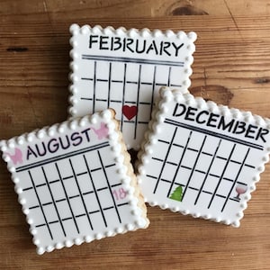 May include: Three calendar-shaped cookies with white icing and black grid lines. The cookies are labeled "FEBRUARY", "AUGUST", and "DECEMBER". Each cookie has a decorative element: a heart, a baby carriage, a Christmas tree, and a wine glass.