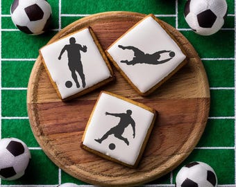 May include: Three white square biscuits decorated with black silhouettes of football players on a wooden board surrounded by white and black footballs.